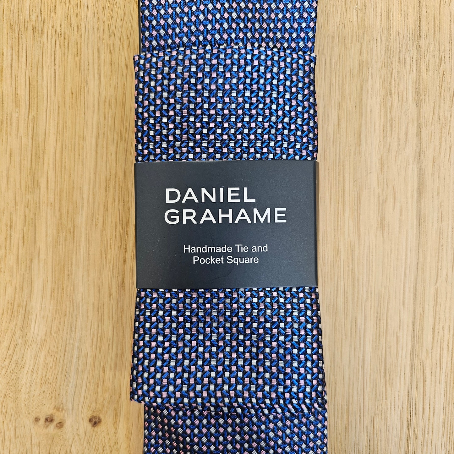 A folded Daniel Grahame necktie with a blue, white, and red micro-pattern, wrapped in a black paper band with white text, displayed on a light wood background.