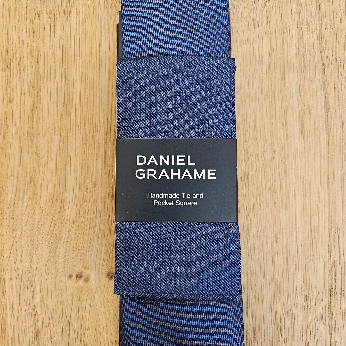 A navy blue silk tie with a textured weave, folded and wrapped in a black paper band featuring the brand name 'DANIEL GRAHAME'. The product is laid flat on a light wood grain surface.