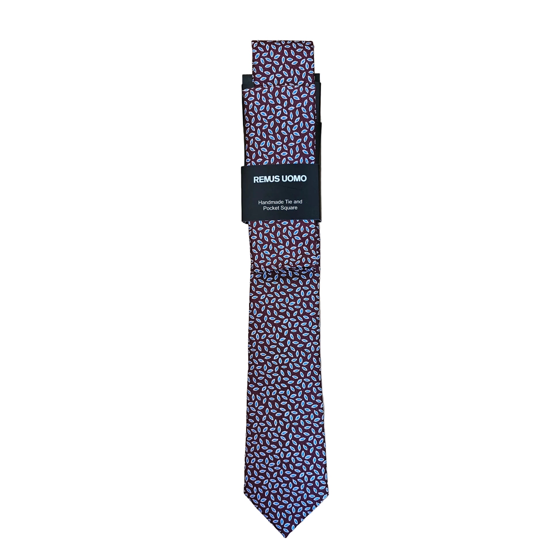 A patterned necktie with a small geometric print in purple, pink, and white, folded with a black paper band that reads 'REMAE LOMBO' on a light wood background.