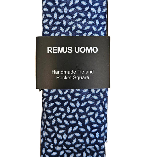 A square navy blue pocket square with a white leaf pattern, wrapped with a black paper band labeled 'REMUS UOMO Handmade Tie and Pocket Square', laid flat on a light wood background.