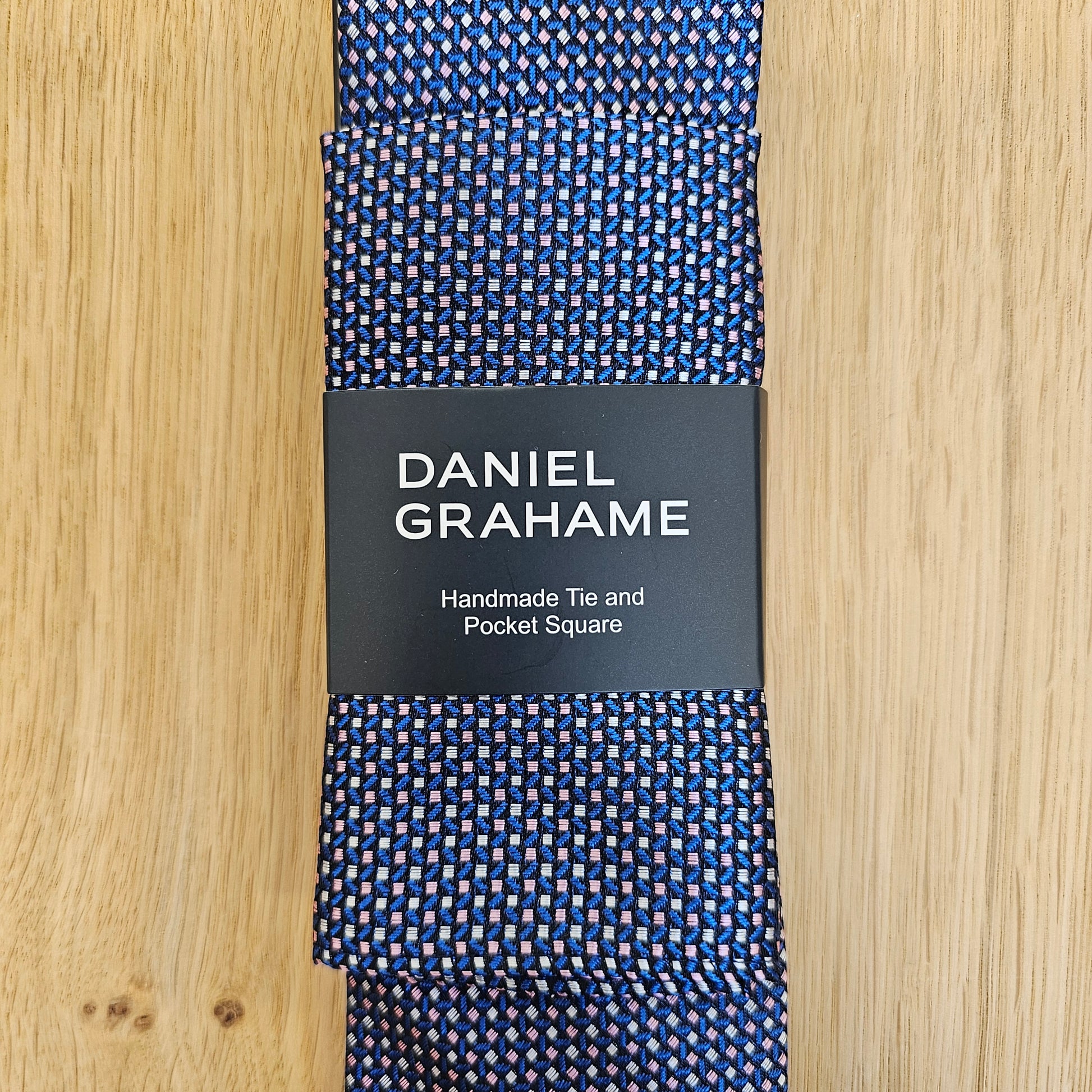 A folded Daniel Grahame necktie with a blue, white, and red micro-pattern, wrapped in a black paper band with white text, displayed on a light wood background.