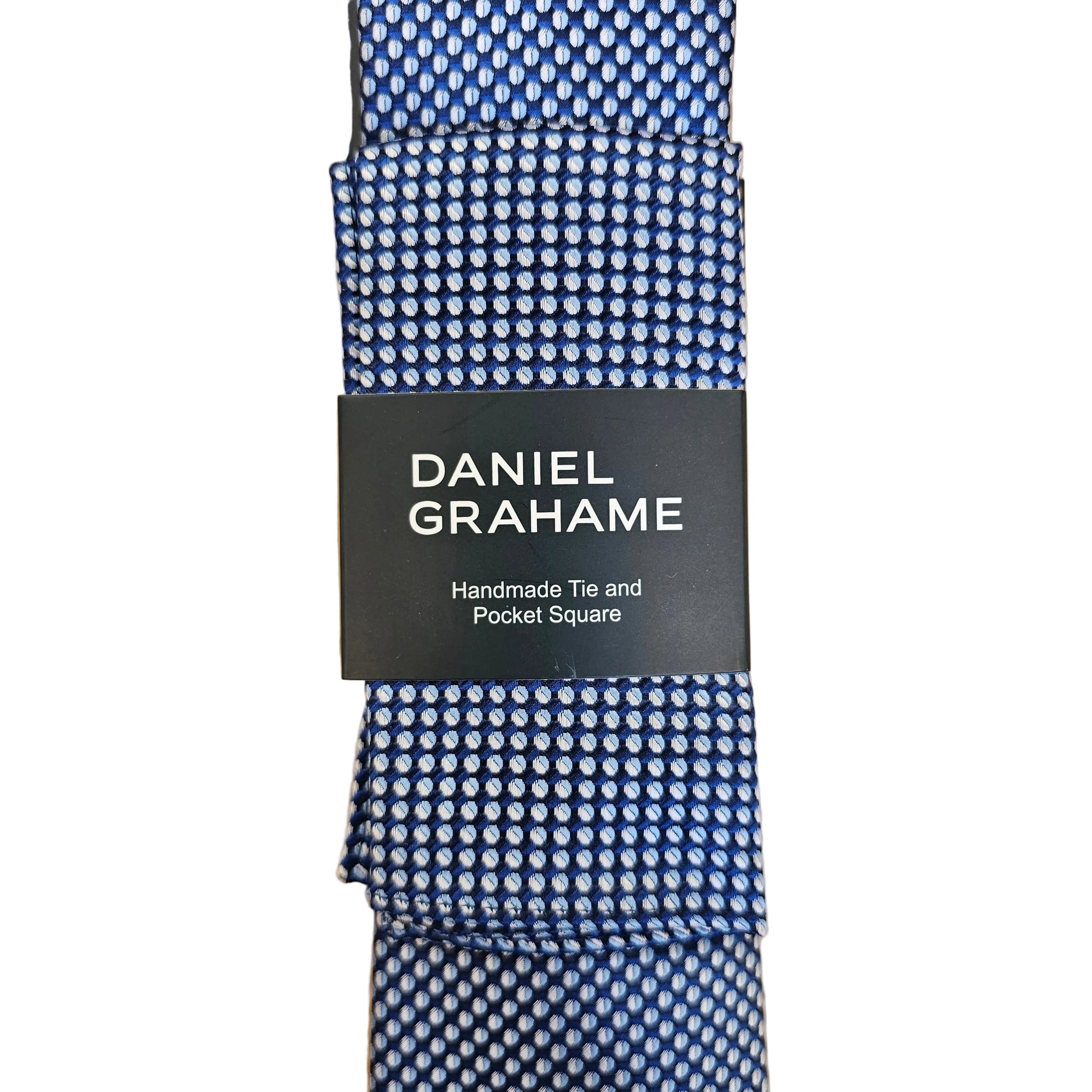 A blue and white patterned necktie with a black paper band label reading 'DANIEL GRAHAME Handmade, Tie and Pocket Square' laid flat on a light wood surface.