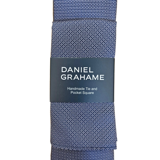 A folded Daniel Grahame tie with a blue and white geometric pattern, wrapped in a dark gray paper band that reads 'DANIEL GRAHAME Handmade Tie and Pocket Square', displayed on a light wood background.