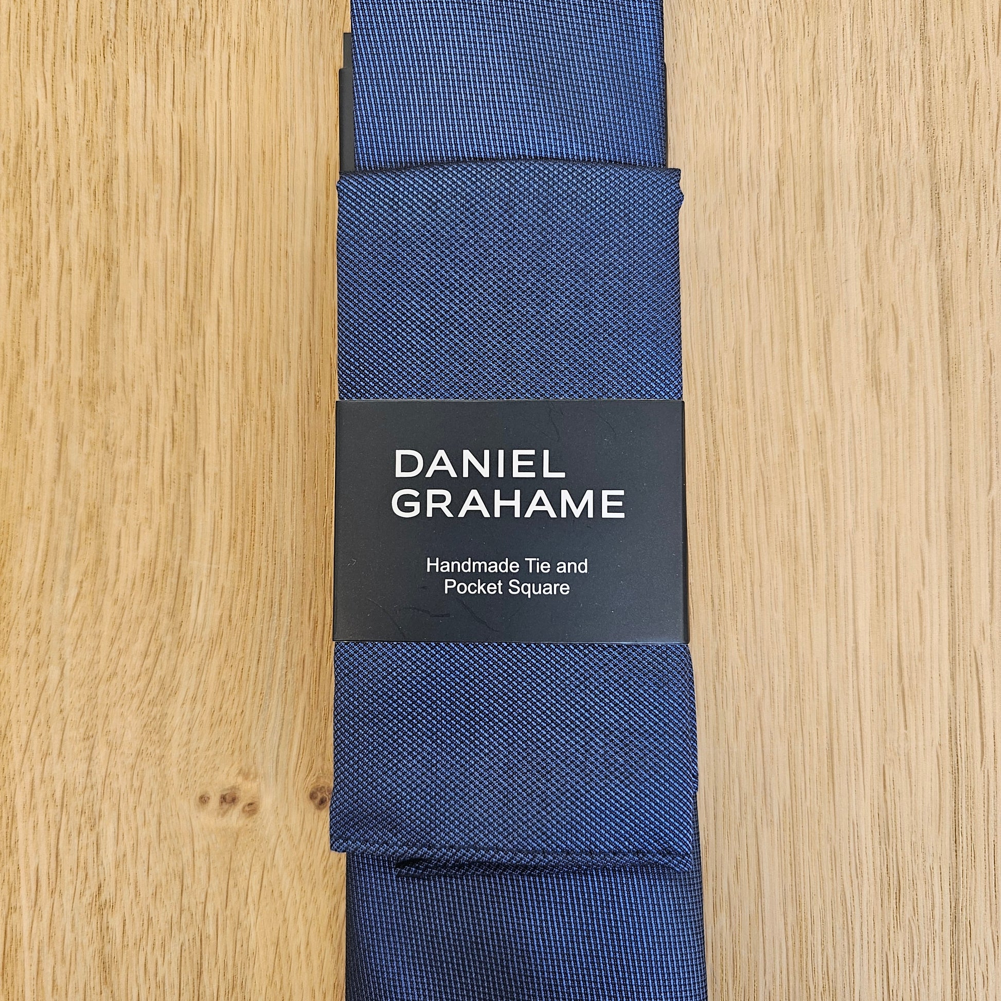 A navy blue silk tie with a textured weave, folded and wrapped in a black paper band featuring the brand name 'DANIEL GRAHAME'. The product is laid flat on a light wood grain surface.