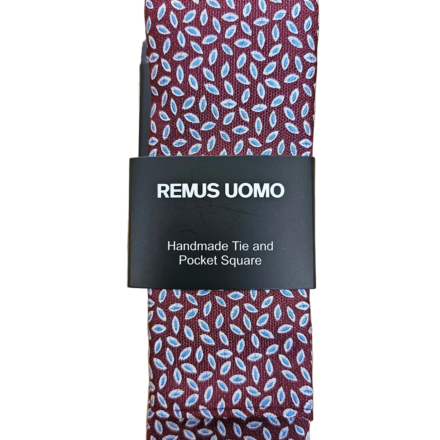 A square pocket square tie with a burgundy base and a light blue and white leaf pattern, wrapped with a black paper band labeled 'REMUS UOMO' on a light wood background.