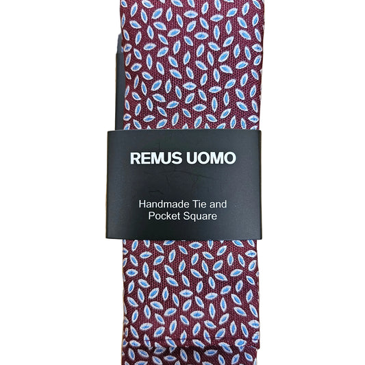 A square pocket square tie with a burgundy base and a light blue and white leaf pattern, wrapped with a black paper band labeled 'REMUS UOMO' on a light wood background.
